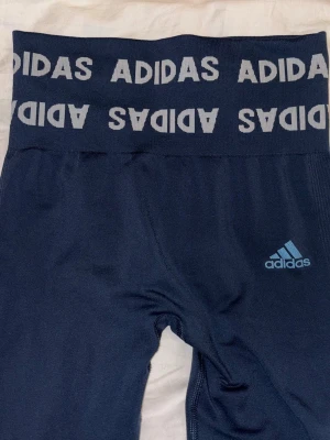 Mörkblå leggings från Adidas - Snygga mörkblå leggings från Adidas med bred resår i midjan där Adidas-loggan upprepas runtom. Tillverkade i stretchigt material som sitter skönt och följsamt. Adidas-logga i ljusblått på ena benet. 