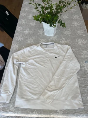 Vit långärmad sweatshirt från Nike - Klassisk vit sweatshirt från Nike med rund halsringning och svart broderad logga på bröstet. Tröjan har långa ärmar och är gjord i mjuk bomull, perfekt för en avslappnad och sportig stil.