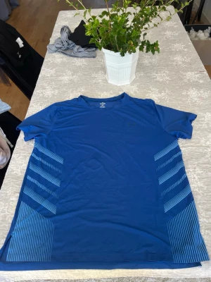 Under armour - Snygg blå träningstopp med korta ärmar och randiga detaljer i ljusblått på sidorna. Tillverkad i ett lätt och snabbtorkande syntetmaterial som passar perfekt för aktiva dagar. Sportig look med diskret logga på bröstet.