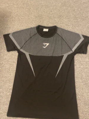 Svart och grå träningströja Gymshark - Snygg svart och grå träningströja från Gymshark med korta ärmar och mönstrade detaljer över axlar och bröst. Tröjan har en sportig passform och är tillverkad i ett lätt och andningsbart material, perfekt för gymmet eller löprundan.