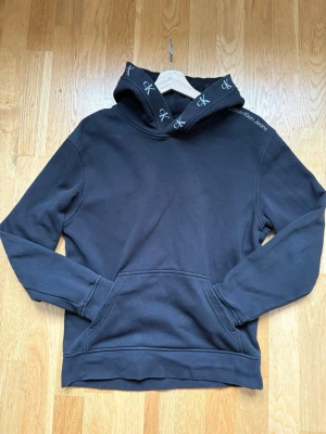 Svart hoodie från Calvin Klein Jeans - Snygg svart hoodie från Calvin Klein Jeans med huva och känguruficka. Huvan har logotypdetaljer med 'ck' och texten Calvin Klein Jeans på axeln. Tillverkad i mjukt material som känns skönt mot huden. Perfekt för en avslappnad och trendig look.