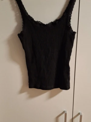 Svart ribbad linnetopp från H&M Divided - Snygg svart ribbad linnetopp från H&M Divided med spetsdetaljer längs ringningen och axelbanden. Perfekt att styla till jeans eller kjol för en trendig look. Tunn och stretchig, passar till många olika outfits.