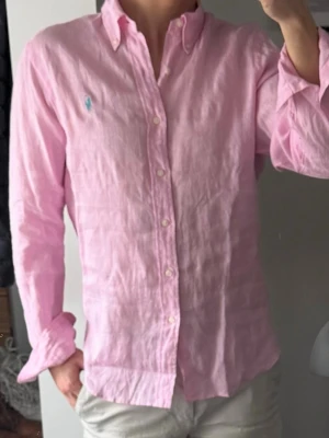 Rosa linneskjorta från Polo Ralph Lauren - Säljer en ljusrosa skjorta från Polo Ralph Lauren i 100% linne med klassisk krage och knäppning framtill. Skjortan har lång ärm och den ikoniska blå logotypen broderad på bröstet. Perfekt för en fräsch och avslappnad stil.