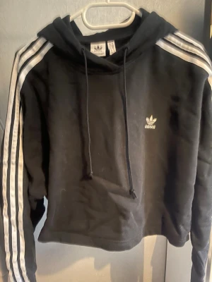 Svart cropped hoodie från Adidas - Svart hoodie från Adidas med klassiska vita ränder längs ärmarna och liten vit logga på bröstet. Croppad modell med huva och dragsnören. Perfekt för en sportig och avslappnad stil. Tillverkad i mjukt material för extra komfort.