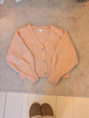 Persikofärgad kofta från H&M - Superfin persikofärgad kofta från H&M med v-ringning och tre dekorativa knappar framtill. Koftan är stickad i ett mjukt material och har ballongärmar för en trendig look. Perfekt att slänga över en topp för en chill stil.