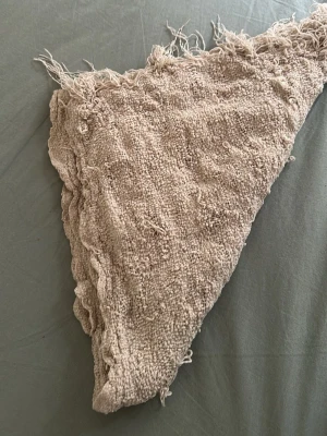 Beige stickad trekantssjal - Mysig beige trekantssjal i grovstickat material med fransar längs ena kanten. Sjalen har en naturlig, bohemisk look och är perfekt att svepa runt halsen för extra värme. Materialet känns mjukt och strukturerat, vilket ger en avslappnad vibe.