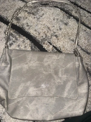 Grå axelväska med kedjedetaljer - Snygg grå axelväska. Väskan har ett lock framtill och ett axelband med silverfärgade kedjedetaljer som ger en cool touch. Kedjan på en av banden har gått av därav ser det annorlunda ut. Perfekt storlek för det viktigaste och enkel att matcha till olika outfits.