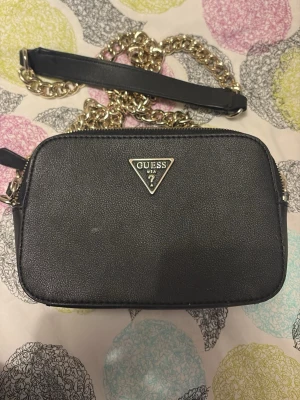 Svart handväska från Guess - Snygg svart handväska från Guess med guldfärgad kedjerem och Guess-logga i metall framtill. Väskan har en rektangulär form och är tillverkad i konstläder. Perfekt för att bära det viktigaste och ger en trendig touch till din outfit.