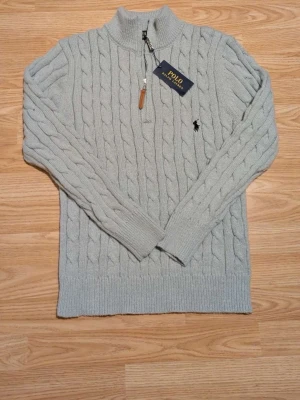 Ljusgrå kabelstickad tröja Polo Ralph Lauren - Snygg ljusgrå kabelstickad tröja från Polo Ralph Lauren med halv dragkedja och hög krage. Tröjan har det klassiska broderade logotypen på bröstet och ribbade muddar vid ärmslut och nederkant. Perfekt för en stilren och avslappnad look.