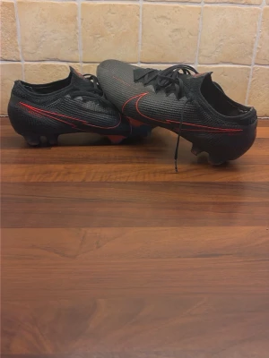 Svarta Nike Mercurial fotbollsskor - Snygga svarta Nike Mercurial fotbollsskor med röda detaljer och klassisk låg profil. Skorna har snörning och är tillverkade i ett lätt syntetmaterial för bra bollkontroll och grepp på planen. Perfekta för dig som vill ha stilrena och snabba skor på matchen.