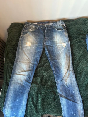 Blå slitna jeans från Replay - Säljer ett par blå jeans från Replay med slitningar och hål på låren för en cool, avslappnad look. Jeansen har klassisk femficksdesign, raka ben och tydliga tvättade detaljer. Perfekta för dig som gillar streetstyle och vintagekänsla.