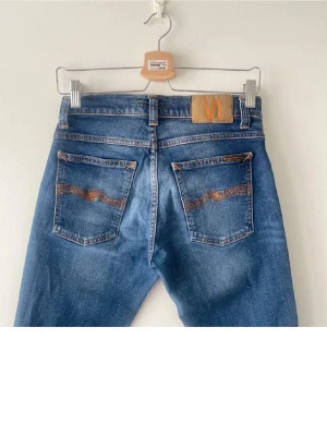 Blå jeans från Nudie Jeans - Säljer ett par klassiska blå jeans från Nudie Jeans med fem fickor och orangea sömmar. Jeansen har raka ben och en snygg tvättad look med Nudie-logga på bakfickan. Perfekta för en avslappnad och trendig stil. Storlek 28/32