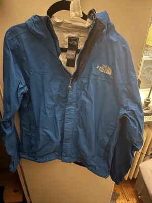 Blå vindjacka från The North Face - Snygg blå vindjacka från The North Face med huva och dragkedja framtill. Jackan har vit logga på bröstet och är tillverkad i lätt, vindtätt material. Perfekt för blåsiga dagar och enkel att packa ner i väskan.