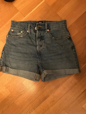 Blå jeansshorts  - Klassiska blå jeansshorts från 157 med uppvikta benslut. Shortsen har fem fickor, bälteshällor och stängs med knapp och dragkedja. Perfekta för en avslappnad stil under varma dagar.