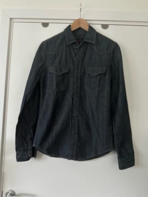 Mörkblå Crocker jeansskjorta med knappar - Mörkblå denim / jeans skjorta från Crocker i storlek XS (för herr, S/M för dam). Klassisk modell med långa ärmar och knäppning framtill och två bröstfickor med lock och knapp. 100% bomull. Mycket bra skick.   Skickas mot porto, kan även hämtas i Stockholm. 