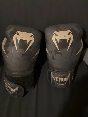 Svarta boxningshandskar från Venum - Säljer ett par svarta boxningshandskar från Venum. Handskarna har kardborreband runt handleden för extra stöd och är tillverkade i slitstarkt syntetmaterial. Perfekta för boxning eller annan kampsportsträning. Original pris 700 och är i nytt skick. Handskarna är 14oz