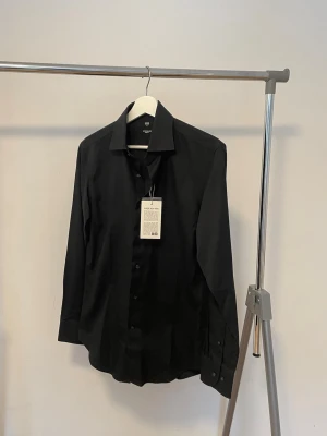 Svart skjorta från Uniqlo - Helt ny aldrig använd svart skjorta från Uniqlo med lång ärm och knappar framtill. Skjortan har en stilren design med krage och är tillverkad i ett mjukt material som känns bekvämt mot huden. Perfekt för en clean och modern look. Pris kan diskuteras vid snabb affär.