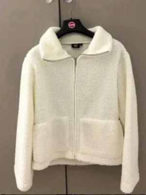 Vit teddyjacka med dragkedja - Säljer en fluffig vit teddyjacka med dragkedja framtill och stora fickor. Jackan har en hög krage och är gjord i mjukt, teddyliknande materia. Helt nytt skick 
