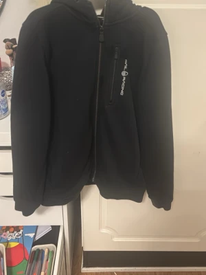 Svart hoodie från Sail Racing - Cool svart hoodie från Sail Racing med dragkedja och huva. Den har en bröstficka med logotyp och är tillverkad i mjukt material som känns skönt mot huden. Perfekt för dig som gillar sportig och stilren design.