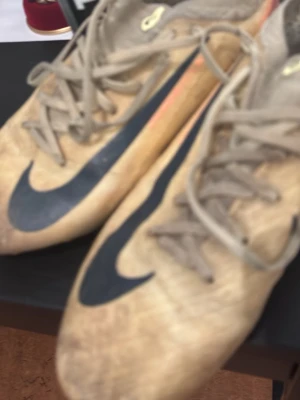 Beige Nike fotbollsskor med dobbar - Säljer ett par beige fotbollsskor från Nike med svarta swoosh-loggor på sidorna. Skorna har snörning och dobbar i orange och svart under sulan, perfekta för spel på gräsplan. Tillverkade i syntetmaterial för bra grepp och känsla på planen.det saknas sula i skon så det får ni köpa själva
