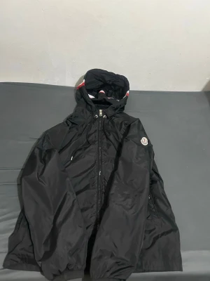 Svart Moncler vindjacka med huva - Snygg svart vindjacka från Moncler med dragkedja framtill och huva med rödvit detalj. Jackan har Moncler-logga på ärmen och två sidofickor med dragkedja. Perfekt för blåsiga dagar och enkel att matcha med olika outfits.