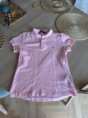 Ljusrosa piké från Ralph Lauren - Säljer en ljusrosa pikétröja från Ralph Lauren med klassisk krage och två knappar framtill. Tröjan har korta ärmar och den ikoniska blå logotypen broderad på bröstet. Perfekt för en avslappnad men ändå stilren look. Riktigt schysst till sommaren