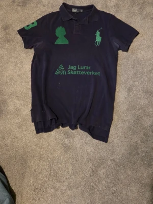 Marinblå piké med grönt tryck - Säljer en marinblå pikétröja från Ralph Lauren med gröna tryck på bröstet och ärmen. Klassisk krage med knappar och korta ärmar. Materialet är mjuk bomull och passformen är normal. Trycket på bröstet har texten 'Jag Lurar Skatteverket'.