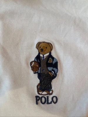 Vit piké Polo Ralph Lauren, polo bear, S - Vit piké T-shirt från Polo Ralph Lauren med klassisk krage, knäppning framtill och broderad björn på bröstet. Polo bear, Teddy. Skjortan är kortärmad och gjord i mjuk bomull för en clean och sportig vibe. Herr S, slim fit.  Köpt på Vinted men passade tyvärr inte mig.  Mindre lagning har gjorts i sprunden på sidorna. Lagningen är välgjord och syns inte vid användning (se bilder). 