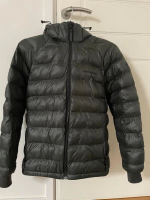 Grön dunjacka från Peak Performance - Snygg grön dunjacka från Peak Performance med huva och svarta dragkedjor. Jackan är quiltad och har en puffig look, perfekt för kalla dagar. Klassisk passform med långa ärmar och praktiska fickor på sidorna.1000 ink frakt kan gå ner lite vid snabb affär. Skick9/10 bara änvänd två månader 