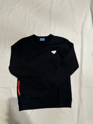 Svart sweatshirt från Prada - Snygg svart sweatshirt från Prada med rund halsringning och ikonisk trekantig logga på bröstet. Tröjan har en röd detalj med text på sidan och är tillverkad i mjukt material för en clean och modern look. Perfekt för dig som gillar exklusiva streetwear-plagg. Pris kan diskuteras 