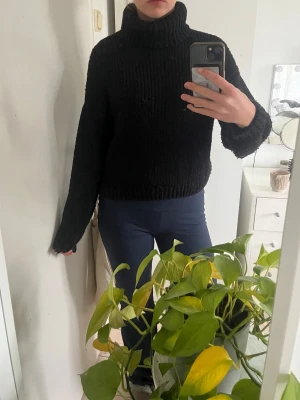 Svart polotröja i grovstickat - Mysig svart polotröja i grovstickat material med lång ärm och hög krage. Tröjan har en avslappnad passform och ribbstickade muddar vid ärmslut och nederkant. Perfekt för kalla dagar och enkel att matcha med olika stilar.