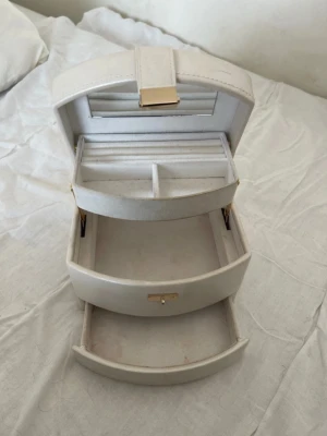 Krämvit smyckeskrin med spegel - Stilren smyckesbox i vit konstläder med guldfärgade detaljer. Boxen har flera fack, utdragbar låda och inbyggd spegel i locket. Perfekt för att organisera ringar, örhängen och halsband. Praktiskt handtag på toppen för enkel transport.