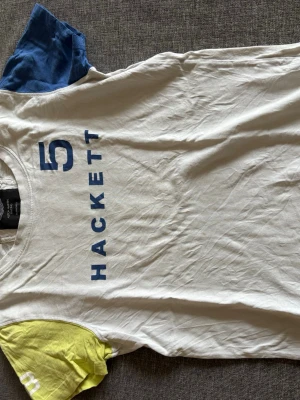 Vit Hackett t-shirt med färgglada ärmar - Vit t-shirt från Hackett x Aston Martin Racing med blått tryck framtill. Ena ärmen är blå och den andra är gul, vilket ger en cool och sportig look. Tillverkad i mjuk bomull och har klassisk passform med rund hals.