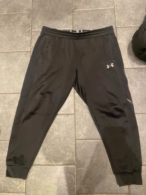Svarta mjukisbyxor från Under Armour - Säljer ett par svarta mjukisbyxor från Under Armour med resår i midjan och muddar vid bensluten. Byxorna har två sidofickor och en diskret logga på låret. Tillverkade i ett mjukt och stretchigt material som passar perfekt för chill eller träning.