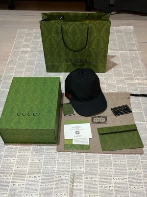 Svart keps från gucci- komplett set - Allt medföljer!!    Snygg svart keps från Gucci med klassisk böjd skärm och ikonisk röd-grön rand på sidan. Kepsen har justerbar rem baktill och är tillverkad i ett slitstarkt tygmaterial. Perfekt accessoar för en trendig och exklusiv look.