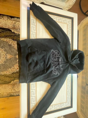 Svart Trapstar Hoodie - Svart hoodie från Trapstar med stor magficka och tryck på bröstet där det står Trapstar Shooters tillsammans med en pantergrafik. Hoodien har dragsko i huvan och är gjord i mjukt bomullsmaterial. Perfekt för en avslappnad streetwear-look.