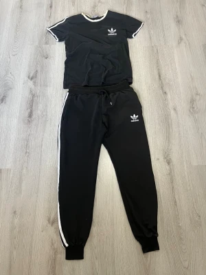 Svart Adidas loungewear-set M - Svart loungewear-set från Adidas med vit kant på t-shirt och vita ränder längs byxorna. Setet består av en kortärmad t-shirt och ett par mjuka byxor med dragsko och muddar. Båda plaggen har Adidas logga framtill. Perfekt för chill dagar hemma. De är barn storlek så de passar folk runt 160 cm