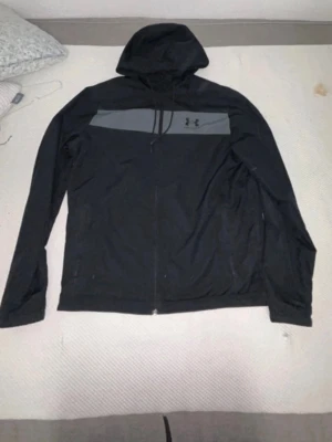 Svart vindjacka från Under Armour - Snygg svart vindjacka från Under Armour med huva och dragkedja framtill. Jackan har en grå panel över bröstet med logga och justerbar huva med snören. Perfekt för blåsiga dagar och sportiga outfits.  Den passar även L Pris kan diskuteras 