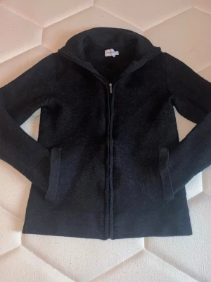 Svart ziptröja i ullblandning - Snygg svart ziptröja med hög krage och två fickor framtill. Tillverkad i mjuk ullblandning som håller dig varm. Perfekt att dra på sig när det är kyligt ute. Enkel och stilren design som passar till det mesta.