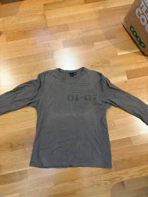 Olivgrön långärmad topp H&M - Olivgrön långärmad topp från H&M med tryckdetalj framtill. Enkel och stilren design med rund halsringning, perfekt att matcha med jeans eller kjol. Tillverkad i mjuk bomull för en skön känsla hela dagen.