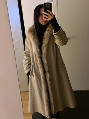 Beige lång kappa med avtagbar päls - Säljer en beige lång kappa med avtagbar päls. Kappan har två snedställda fickor och justerbara ärmslut. Perfekt för kalla dagar och ger en lyxig känsla till din outfit.
