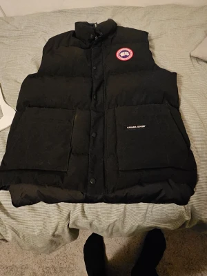 Svart dunväst från Canada Goose - Snygg svart dunväst från Canada Goose med klassisk logga på bröstet och stora fickor framtill. Västen har hög krage, tryckknappar och är tillverkad i ett slitstarkt material med dunfyllning. Perfekt för lager-på-lager-stil under kyliga dagar. Använd en gång typ sen försvann den in i garderoben