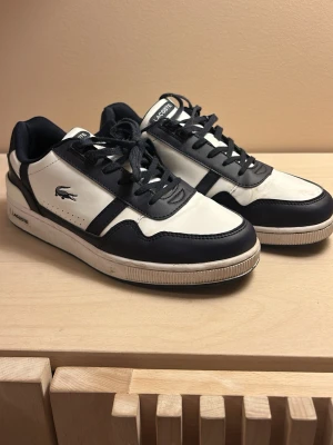 Svarta och vita Lacoste sneakers - Snygga sneakers från Lacoste i svart och vitt läder med klassisk krokodil-logga på sidan. Skorna har svarta snören, vit sula och låg profil. Perfekta för dig som gillar stilrena och sportiga skor med en tidlös vibe.