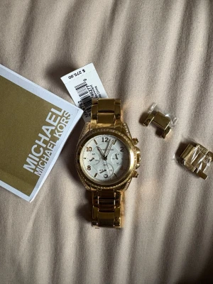 Guldig klocka från Michael Kors - Snygg guldig klocka från Michael Kors med vit urtavla och kristalldetaljer runt boetten. Klockan har länkarmband i metall och flera små urtavlor för extra funktioner. Perfekt accessoar för dig som vill sticka ut lite extra.