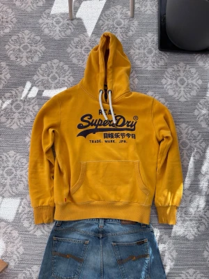 hoodie från Superdry - Säljer en gul hoodie från Superdry med stor logga och japansk text på bröstet. Hoodien har vit dragsko i huvan och en klassisk känguruficka framtill. Tillverkad i mjukt bomullsmaterial och har ribbade muddar vid ärmar och nederkant.