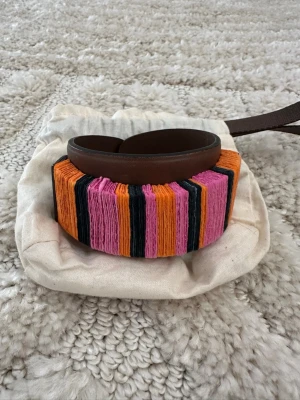 Färgglatt armband från Marni - Unikt armband från Marni i brunt läder med inslag av färgglada trådar i orange, rosa och svart. Bred design som ger ett statement till din outfit. Perfekt för dig som gillar att sticka ut med accessoarer och vill ha något som känns både trendigt och lekfullt.