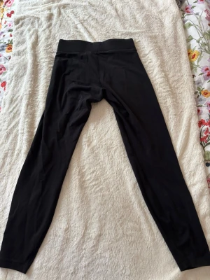 Svarta leggings från Nike - Säljer ett par klassiska svarta leggings från Nike med diskret logga vid benet. De har hög midja och är tillverkade i ett stretchigt material som sitter skönt och följer kroppen. Perfekta till träning eller chill. Passar dig som gillar enkel och sportig stil.