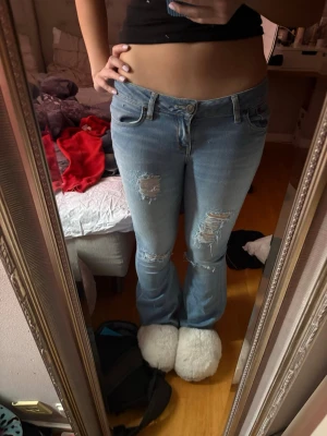 Ljusblå slitna bootcut jeans - Säljer ett par ljusblå bootcut jeans med slitningar och hål på båda benen. Jeansen har låg midja och klassisk femficksdesign. Perfekta för en avslappnad och trendig look. Materialet är denim i bomull.