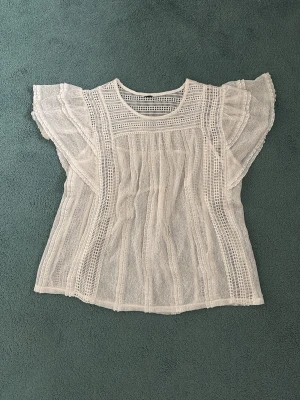 Vit spetsblus med volangärmar - Säljer en superfin vit blus i spets med genombrutna detaljer och volangiga korta ärmar. Blusen har rund halsringning och ett luftigt, lätt material som ger en romantisk vibe. Perfekt att styla med jeans eller kjol för en trendig look.