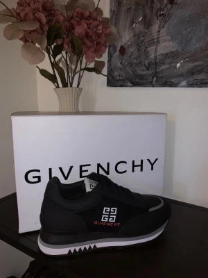 Svarta Givenchy sneakers med logga - Stilrena svarta sneakers från Givenchy med vit och röd logga på sidan. Skorna har en låg siluett, meshdetaljer och inslag av läder. Sulan är vit med svarta detaljer och har ett sågtandat mönster. Perfekta för dig som vill ha en exklusiv och trendig look.